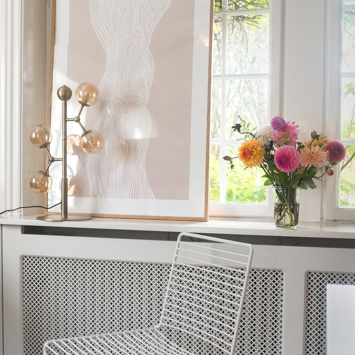 Atom Glass 4 Light Table Lamp in antique brass with amber glass spheres branching from a slim stem, shown on a windowsill beside artwork and fresh flowers.