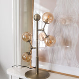 Atom Glass 4 Light Table Lamp in antique brass with amber glass spheres branching from a slim stem, shown on a white console beside framed wall art.