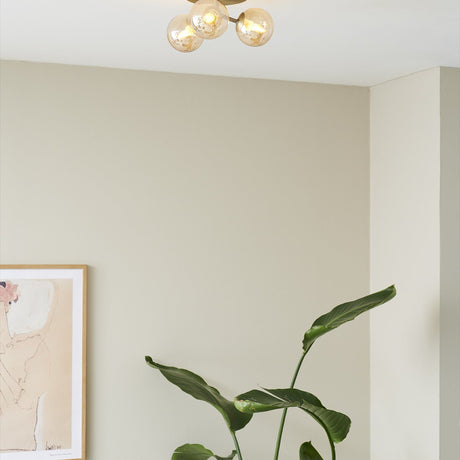 Atom Trio 3 Light Flush Ceiling Light in antique brass with three amber glass spheres, mounted on a light ceiling above a room with a plant and framed art.