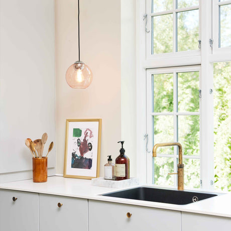 Bubbles Pendant Light 18cm in Rose hangs above a kitchen counter, its dusty pink glass globe on a black cap and cord, with an exposed bulb visible through the clear-tinted glass.