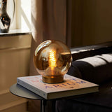 Bubbles Table Lamp 18cm in amber glass with a spherical shade on a low brass base, glowing coil bulb visible, styled on a book beside a sofa.