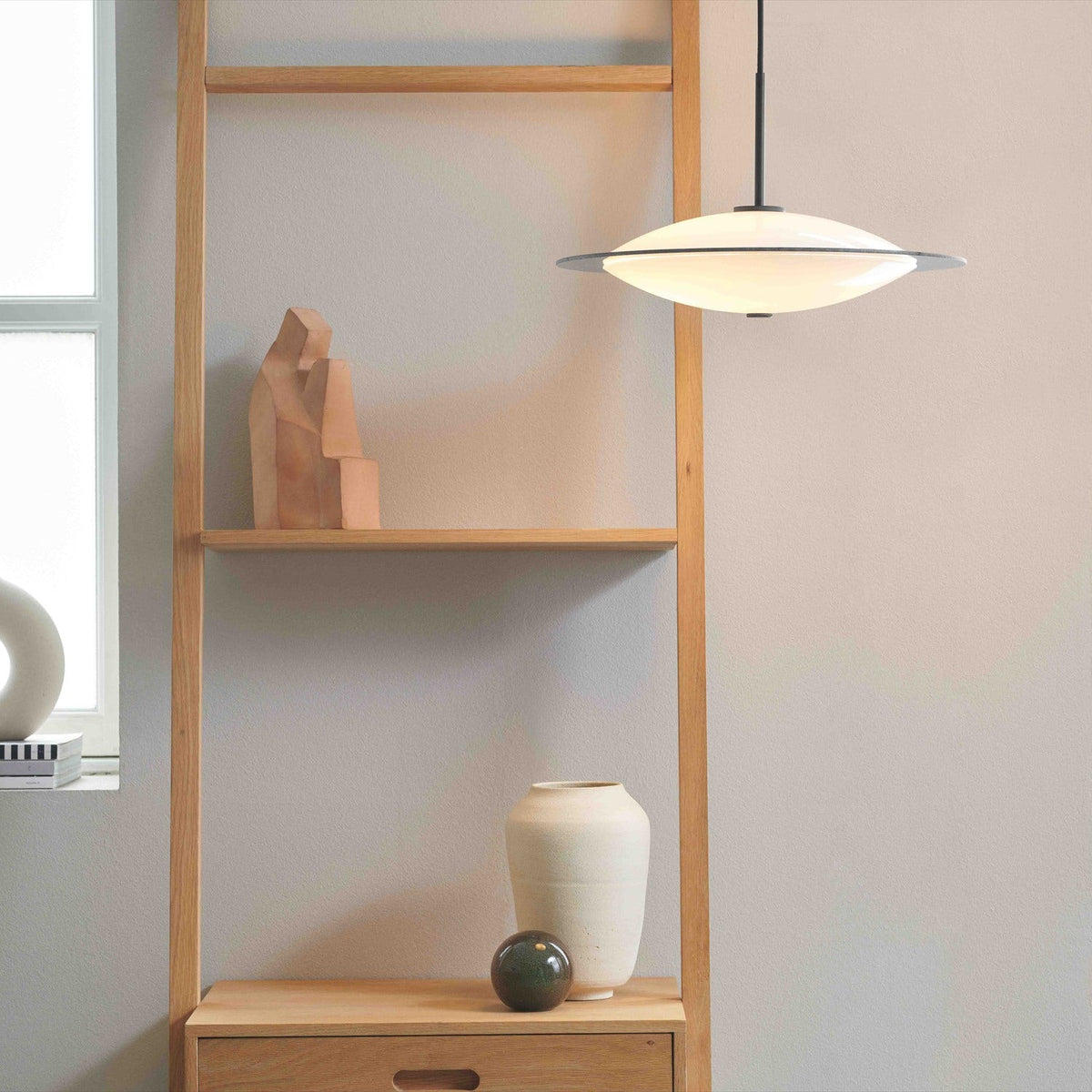 Orbit Glass 3 Light Pendant Light 40cm in opal and black, oval flattened glass shade with a wide flat black disc rim, hung above a wooden shelving unit in a living space.