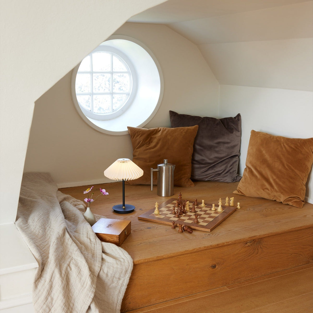 Paris LED Go Clean rechargeable table lamp with white pleated paper shade and slim black stem, shown lit on a wooden bench beside a chess set in a cosy attic nook.