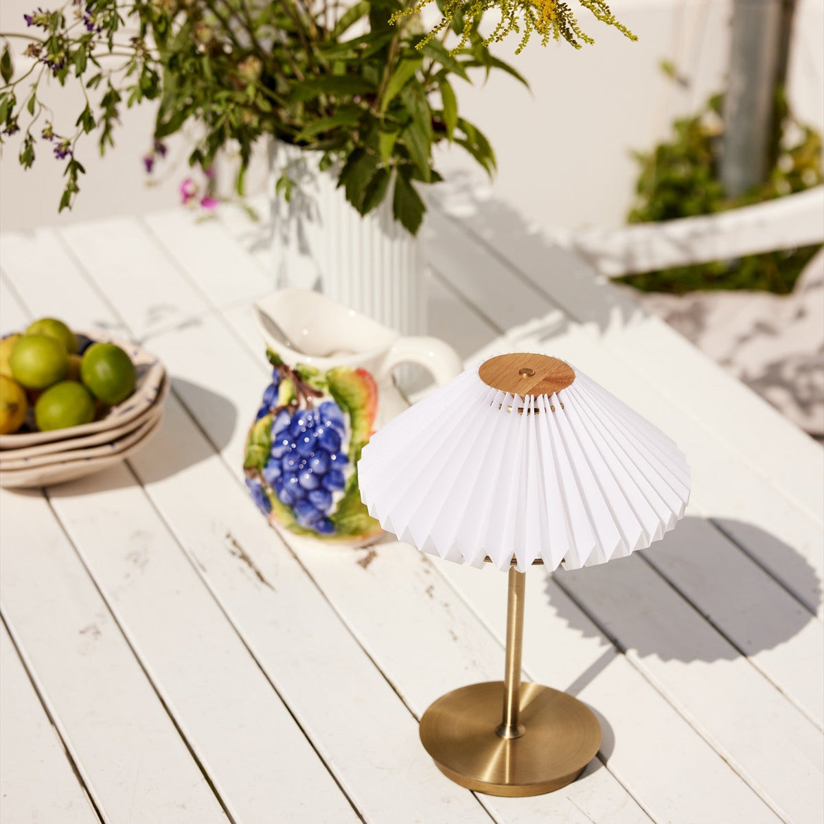 Paris LED Go Rechargeable Table Lamp 22cm in white and antique brass, with pleated shade on a slim brass stem, shown outdoors on a white table beside a fruit bowl and jug.
