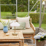 Paris LED Go rechargeable table lamp with pleated white shade and antique brass stem, shown on a rattan coffee table in a bright conservatory seating area.