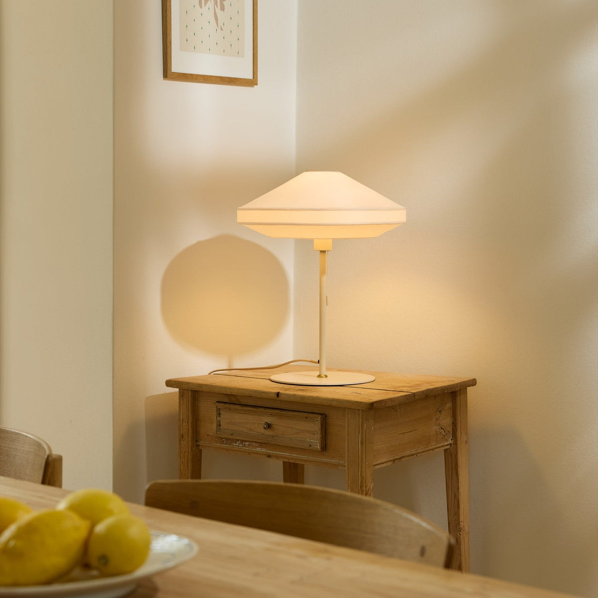 Paris Silk Table Lamp in natural cream fabric with a stepped conical shade on a slim stem and round base, lit on a wooden side table.