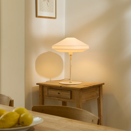 Paris Silk Table Lamp in natural cream fabric with a stepped conical shade on a slim stem and round base, lit on a wooden side table.