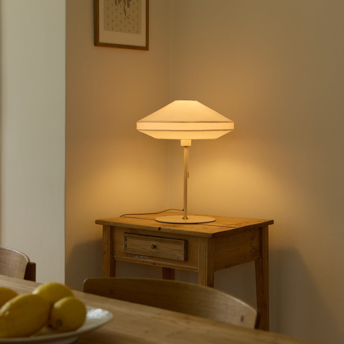 Paris Silk Table Lamp in natural cream fabric with a two-tier shade, slim stem and round base, lit on a wooden side table beside a dining setting.