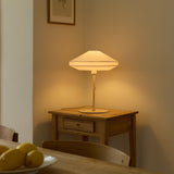 Paris Silk Table Lamp in natural cream fabric with a two-tier shade, slim stem and round base, lit on a wooden side table beside a dining setting.