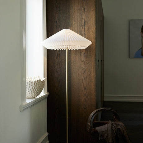 Paris Clean Floor Lamp 56cm in white pleated paper shade with pointed rim on a slim antique brass stem, shown against a dark wood panel in a living room.