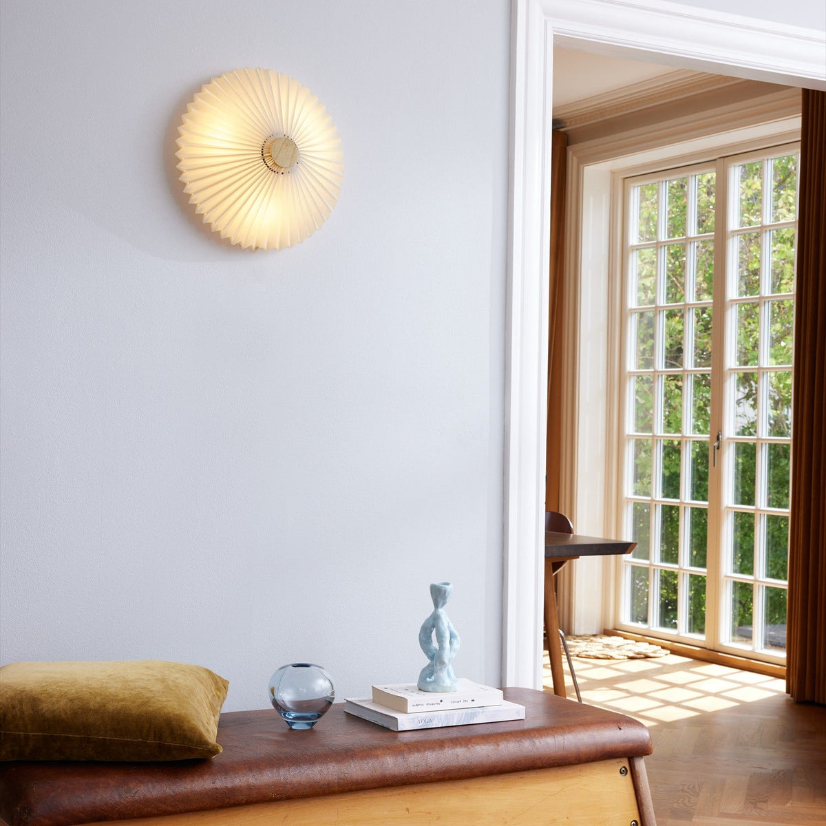 Paris Clean Flush Ceiling Light 50cm illuminated on a living room wall, its white pleated paper shade with pointed edge and antique brass centre casting a warm glow.