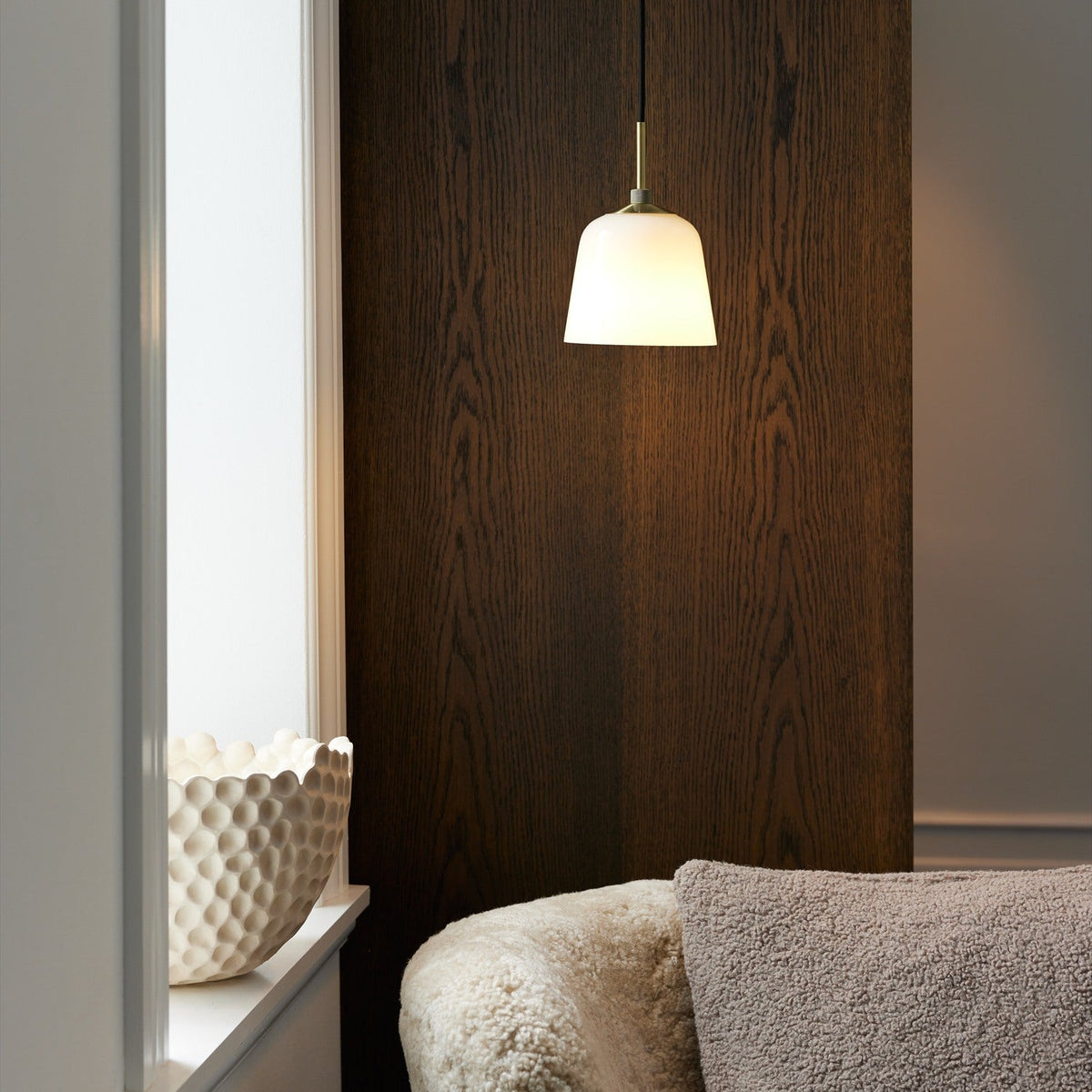 Room Pendant Light 15cm in opal glass with antique brass collar, shown lit above a window sill in a living room with wood panelling and a cream armchair.
