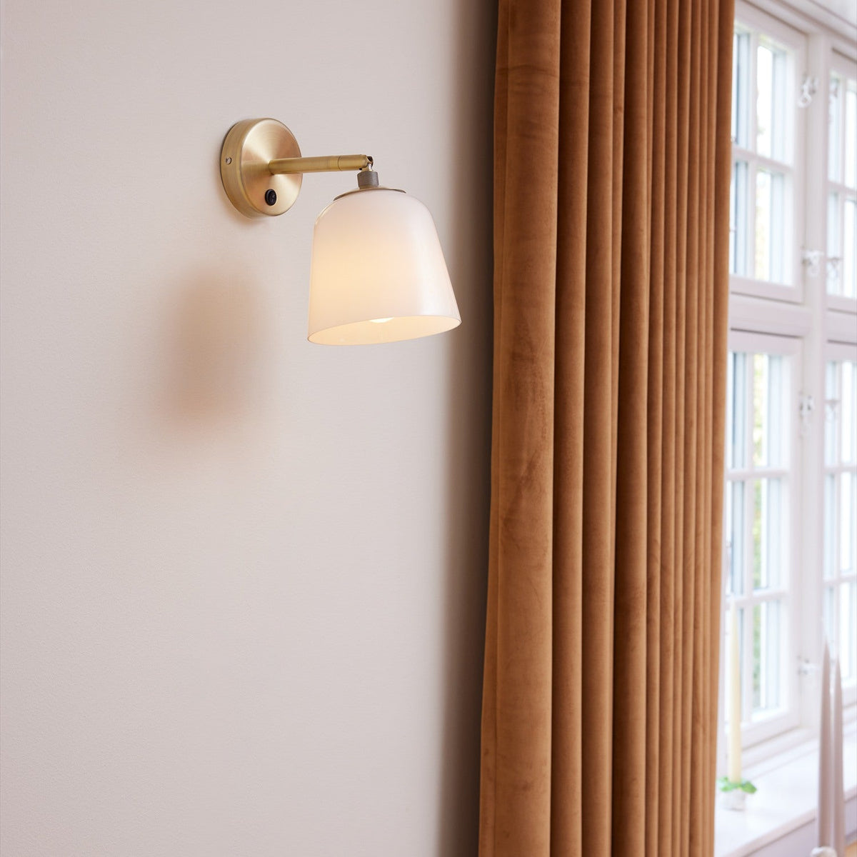 Room Wall Light in opal glass and antique brass, jointed adjustable arm on a round wall plate with switch, shown beside a curtained window in a living room setting.