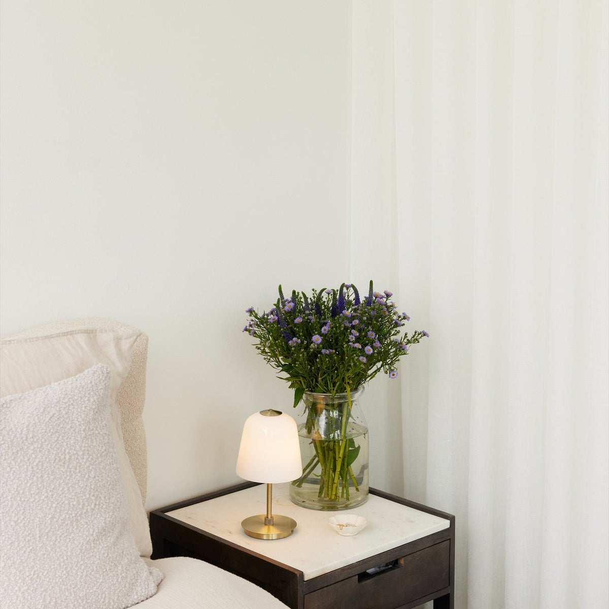 Room LED table lamp with opal bell-shaped glass shade and antique brass stem and base, lit on a marble-top bedside table beside a vase of purple flowers.