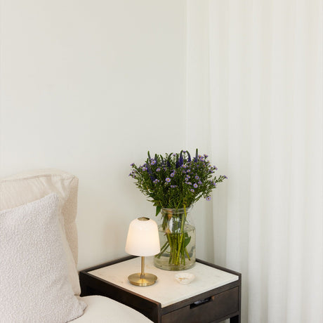 Room LED table lamp with opal bell-shaped glass shade and antique brass stem and base, lit on a marble-top bedside table beside a vase of purple flowers.