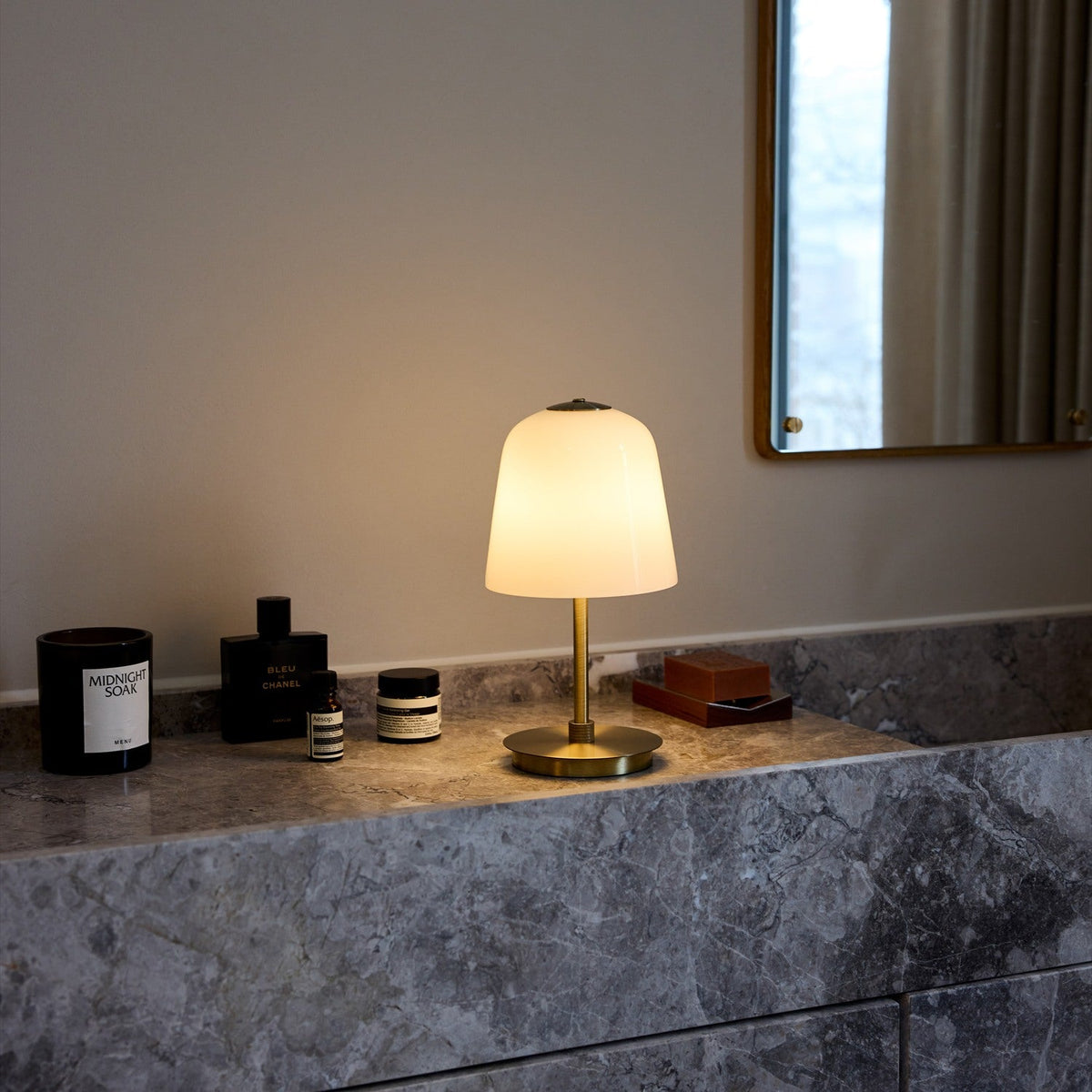 Room LED table lamp with tall bell-shaped opal glass shade on a slim antique brass stem and round base, lit on a marble bathroom countertop beside candles and toiletries.