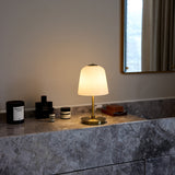 Room LED table lamp with tall bell-shaped opal glass shade on a slim antique brass stem and round base, lit on a marble bathroom countertop beside candles and toiletries.