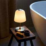 Room LED 2.5W Outdoor Table Lamp in opal and antique brass, bell-shaped glowing glass shade on a slim brass stem with round base, shown lit on a wooden stool beside a bathtub.