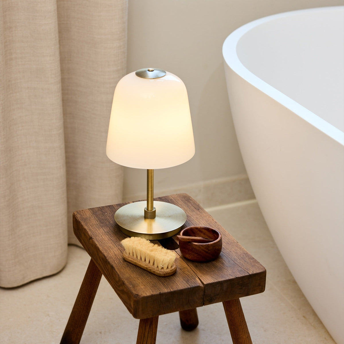 Room LED table lamp with opal glass bell shade and antique brass stem and base, lit on a wooden stool beside a bathtub.