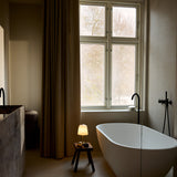 Room LED table lamp with opal glass bell shade and brass stem, glowing softly on a wooden stool beside a freestanding bathtub.