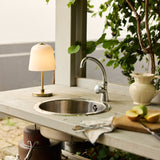Room LED Outdoor Table Lamp with opal bell-shaped glass shade and antique brass base, lit on an outdoor kitchen counter beside a sink and jug.