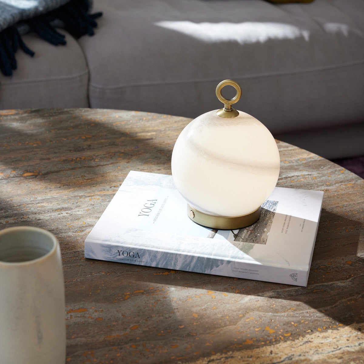 Stockholm LED Rechargeable Outdoor Table Lamp 15cm in Marble Grey, a spherical opal glass globe with grey-brown marbling on a brass base and ring handle, shown on a book atop a wooden coffee table.
