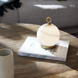 Stockholm LED Rechargeable Outdoor Table Lamp 15cm in Marble Grey, a spherical opal glass globe with grey-brown marbling on a brass base and ring handle, shown on a book atop a wooden coffee table.