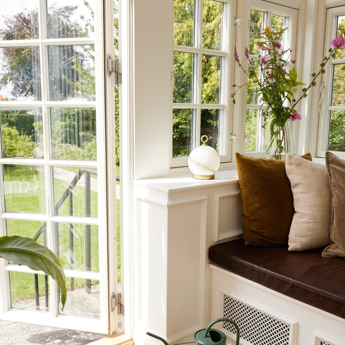 Stockholm LED Rechargeable Outdoor Table Lamp 15cm in marble green, opal glass globe in a brass cup with brass ring handle, shown on a windowsill beside cushions and flowers.