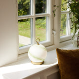 Stockholm LED Rechargeable Outdoor Table Lamp 15cm in Marble Green, an opal glass globe with green marbled streaks on a brass base and ring handle, shown on a windowsill.