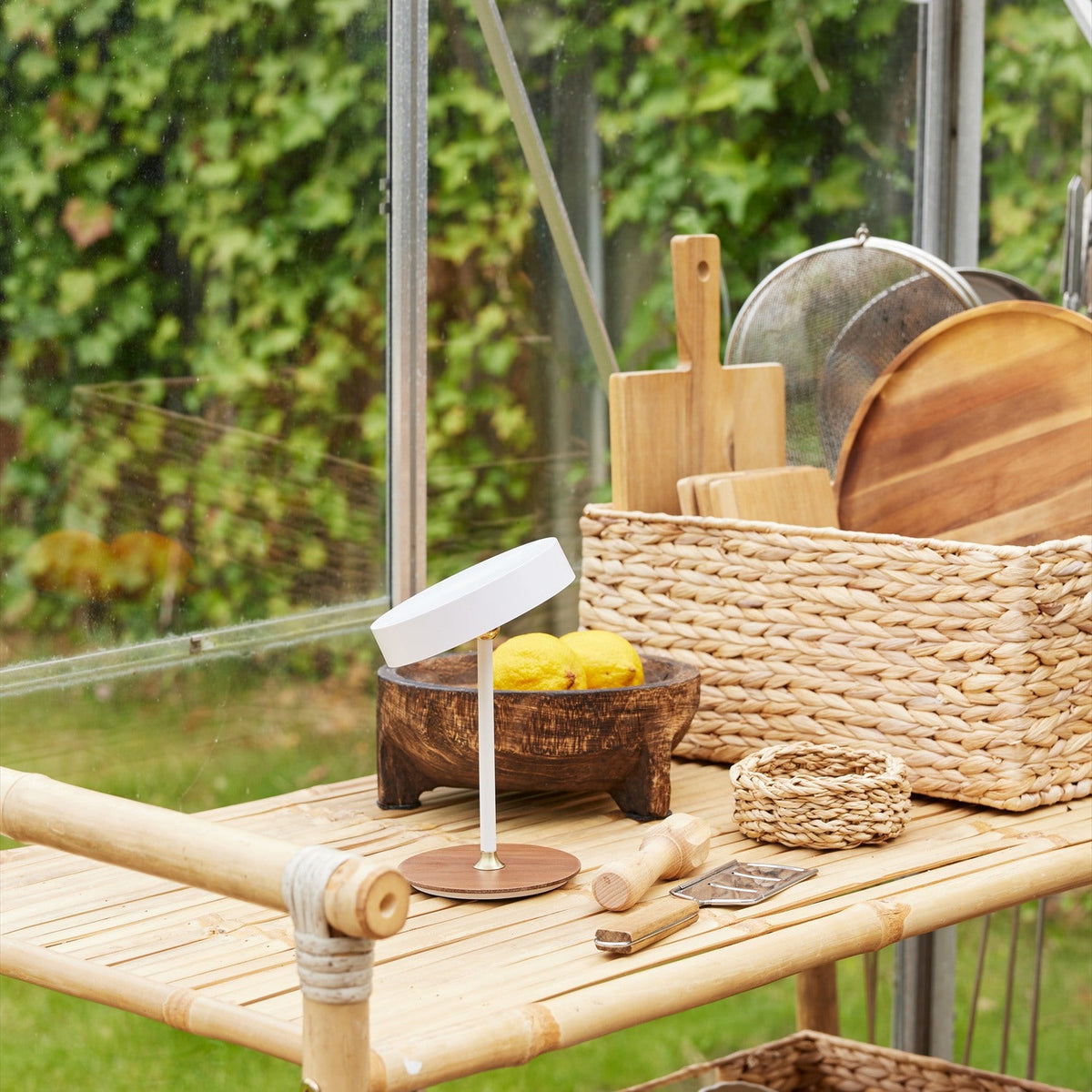 On the Move LED Rechargeable Table Lamp 13cm in white with a round walnut base and brass collar, shown on a bamboo shelf in a garden setting.