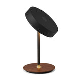 On the Move LED rechargeable table lamp in black, tilted round drum head on a slim black stem with brass collar, set on a walnut wood disc base, plain white background.