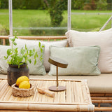 On the Move LED Rechargeable Table Lamp 13cm in antique brass with a round wood base, shown on a bamboo table beside cushions, lemons, and a potted mint plant.