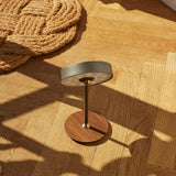 On the Move LED rechargeable table lamp, 13cm, gunmetal drum shade on a slim stem with brass collar and round walnut base, shown on a wooden floor beside a woven jute rug.