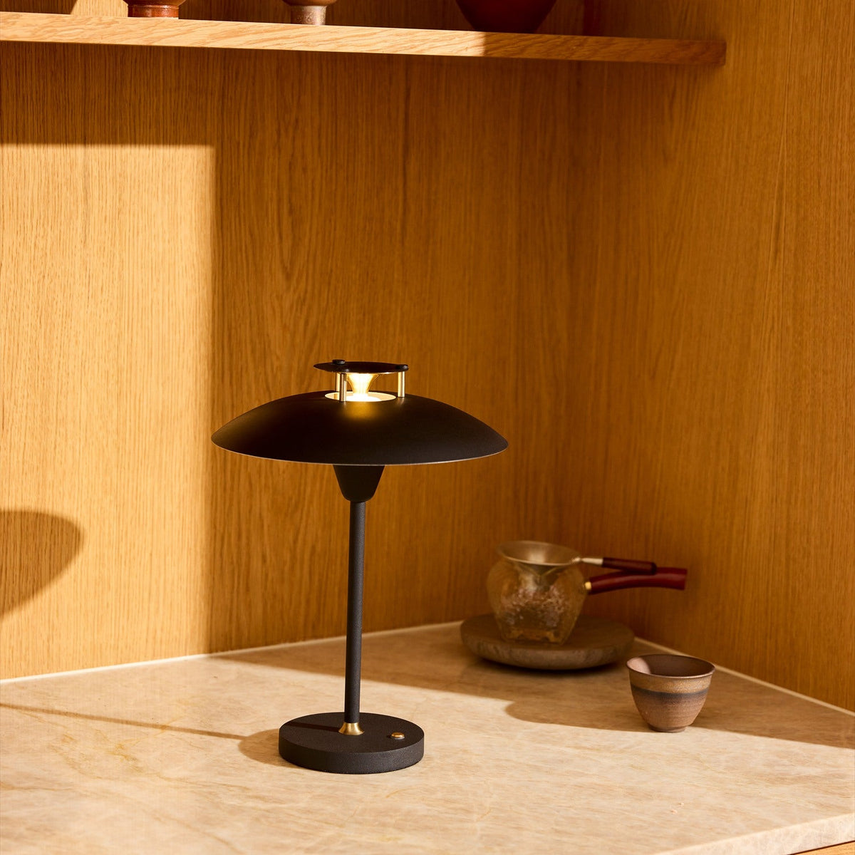 Stepp LED Outdoor Table Lamp in black with a wide flat disc shade and brass accents, shown lit on a stone surface against a wood-panelled wall.