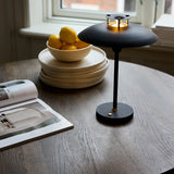 Stepp LED Outdoor Table Lamp in black, wide flat disc shade with brass detailing atop a slim stem and round base, shown lit on a wooden table beside a bowl of lemons.