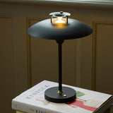Stepp LED Outdoor Table Lamp in black, wide flat disc shade with stepped brass-accented cap glowing warm light, on a round black base atop a stacked books display.