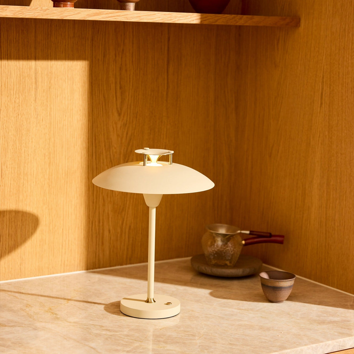 Stepp LED Outdoor Table Lamp in cream with a wide flat disc shade and slim stem on a round base, shown lit on a marble surface against a wooden interior.