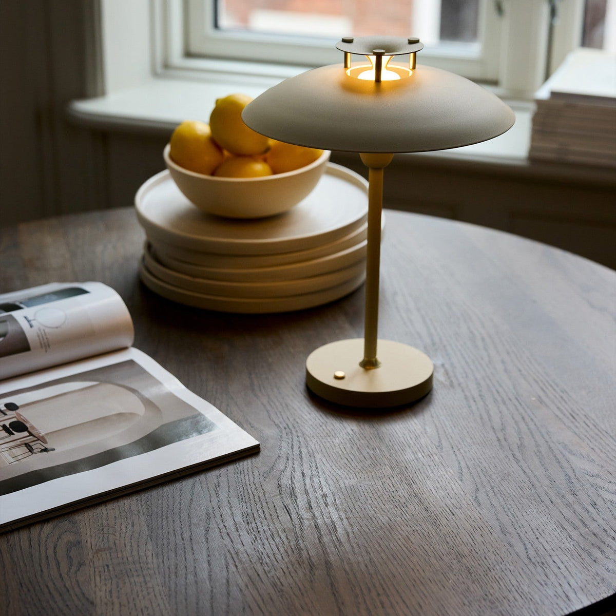 Stepp LED Outdoor Table Lamp in cream, with a wide flat disc shade and slim stem on a round base, shown lit on a wooden table beside a fruit bowl and magazine.