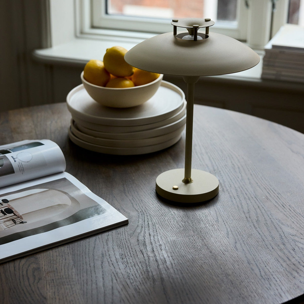 Stepp LED Outdoor Table Lamp in cream, wide flat disc shade on a slim stem with round base, shown on a wooden table beside a bowl of lemons and plates.