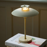 Stepp LED Outdoor Table Lamp in cream with a wide flat disc shade, stepped cap, slim stem and round base, glowing warmly on a book beside a panelled wall.