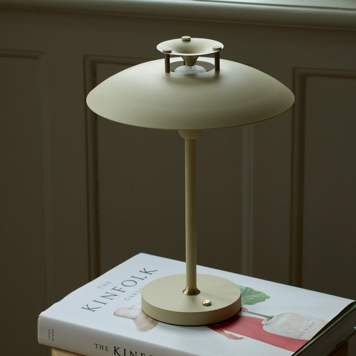 Stepp LED Outdoor Table Lamp in cream, with a wide flat disc shade, slim stem and round base with brass button, shown glowing on a book beside a panelled wall.