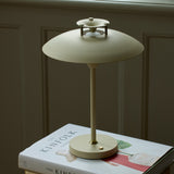 Stepp LED Outdoor Table Lamp in cream, with a wide flat disc shade, slim stem and round base with brass button, shown glowing on a book beside a panelled wall.
