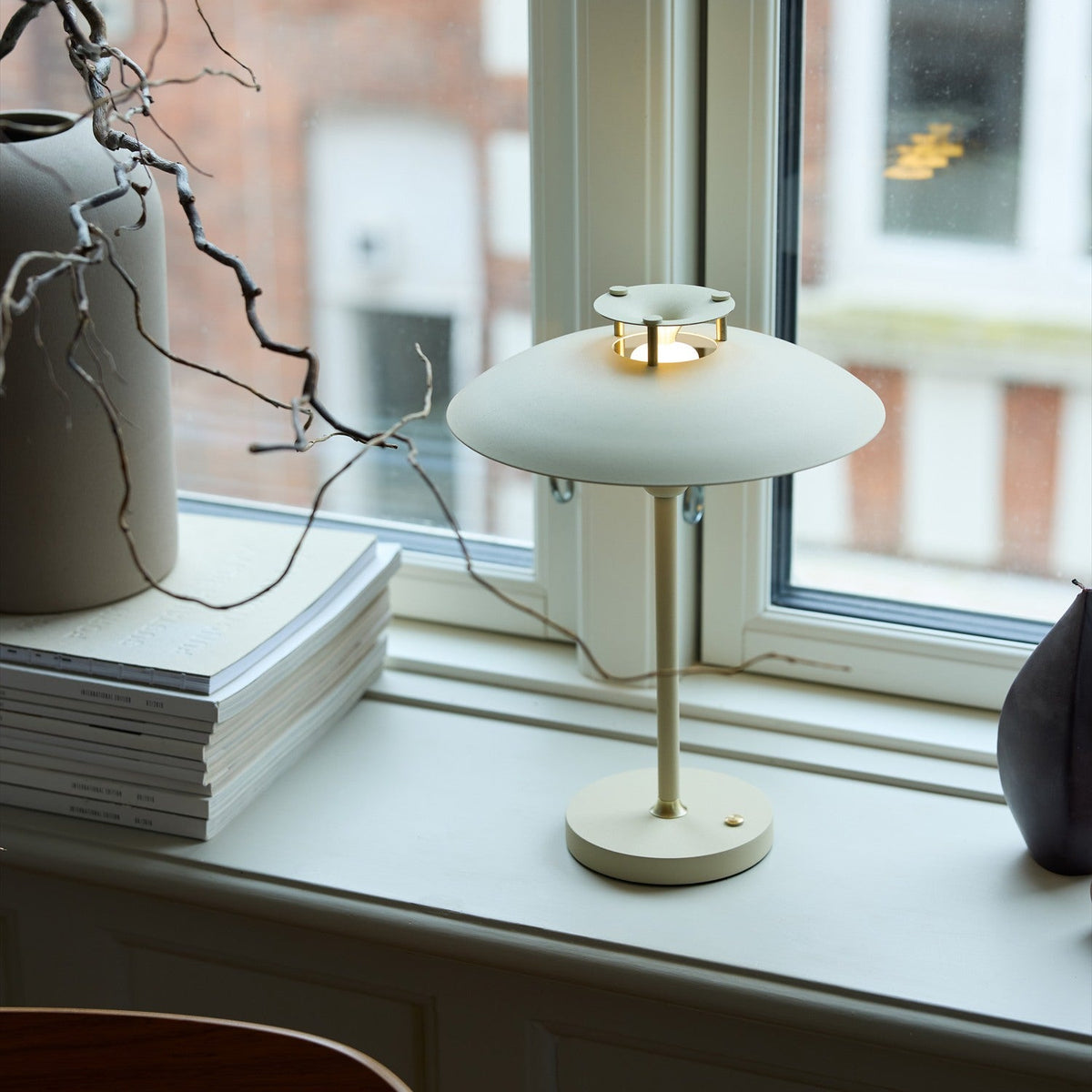 Stepp LED Outdoor Table Lamp in cream, with a wide flat disc shade and slim stem on a round base, glowing softly on a windowsill beside books and a vase.