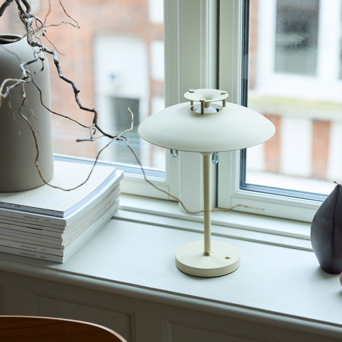 Stepp LED Outdoor Table Lamp in cream, with a flat disc shade and slim stem on a round base, shown on a windowsill beside books and a vase.