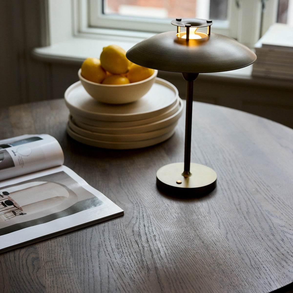 Stepp LED Outdoor Table Lamp in antique brass with a wide shallow disc shade and slim stem, lit on a wooden table beside a lemon bowl and stacked plates.