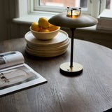 Stepp LED Outdoor Table Lamp in antique brass with a wide shallow disc shade and slim stem, lit on a wooden table beside a lemon bowl and stacked plates.