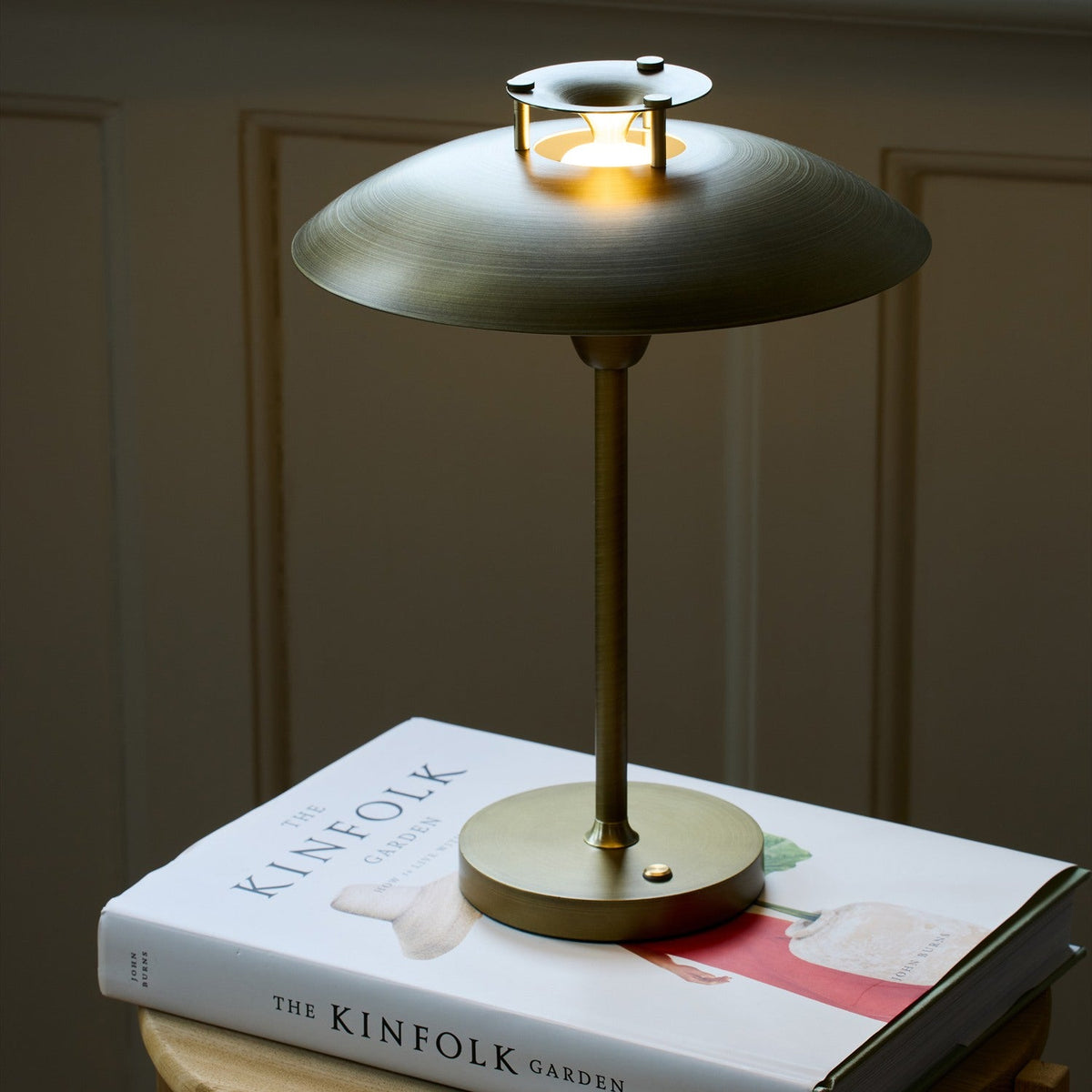 Stepp LED outdoor table lamp in antique brass with a wide flat disc shade glowing beneath a stepped cap, slim stem and round base, shown lit atop a book indoors.