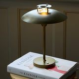 Stepp LED outdoor table lamp in antique brass with a wide flat disc shade glowing beneath a stepped cap, slim stem and round base, shown lit atop a book indoors.