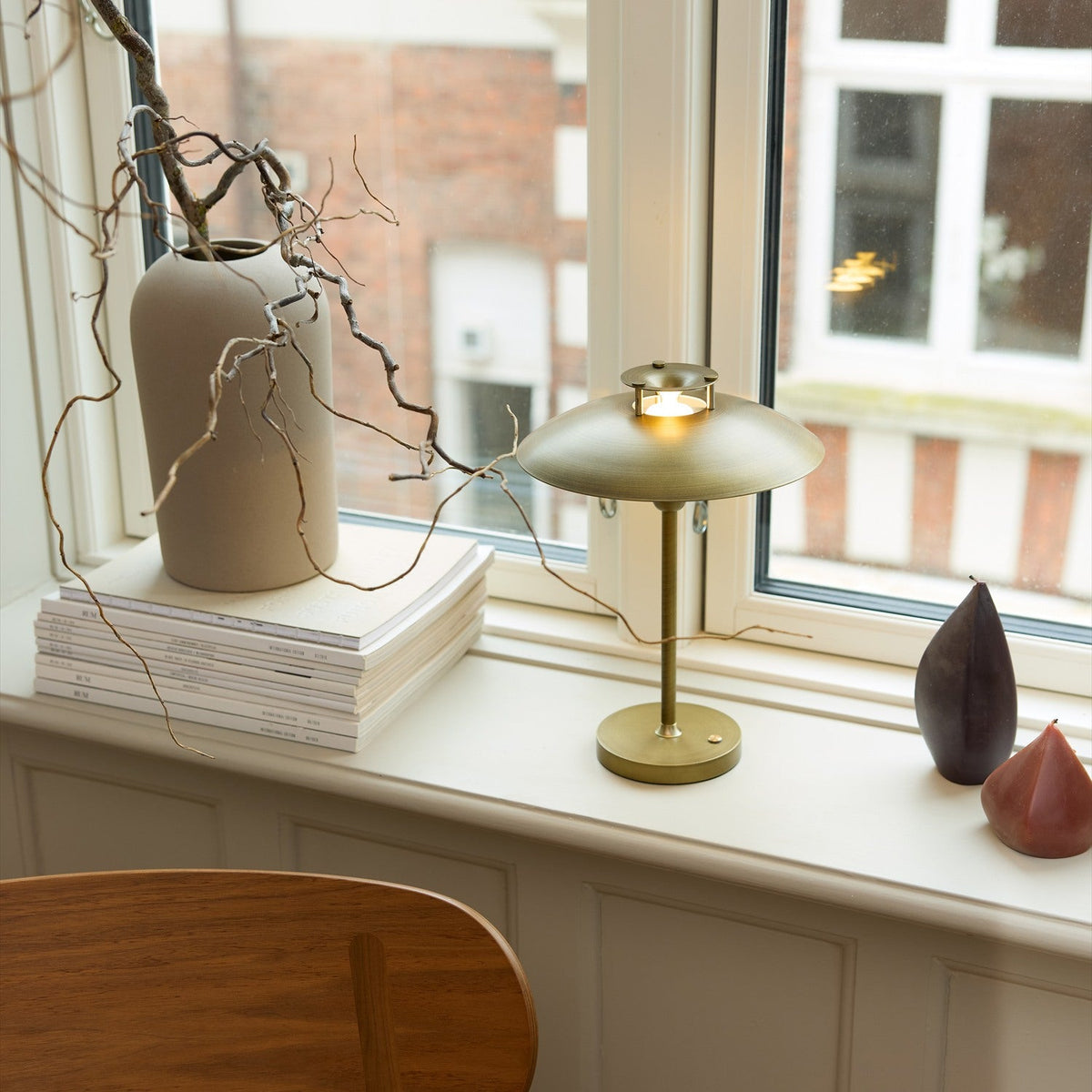 Stepp LED Outdoor Table Lamp in antique brass with a wide flat disc shade and slim stem, shown lit on a windowsill beside a vase and books.
