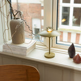 Stepp LED Outdoor Table Lamp in antique brass with a wide flat disc shade and slim stem, shown lit on a windowsill beside a vase and books.